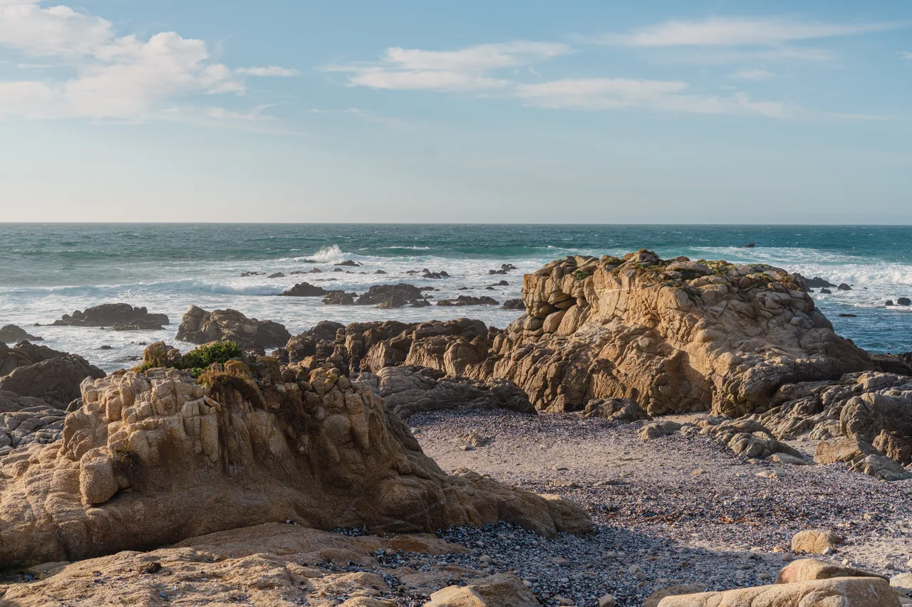 Sunlit coastal rocks beside breaking waves and a blue horizon.